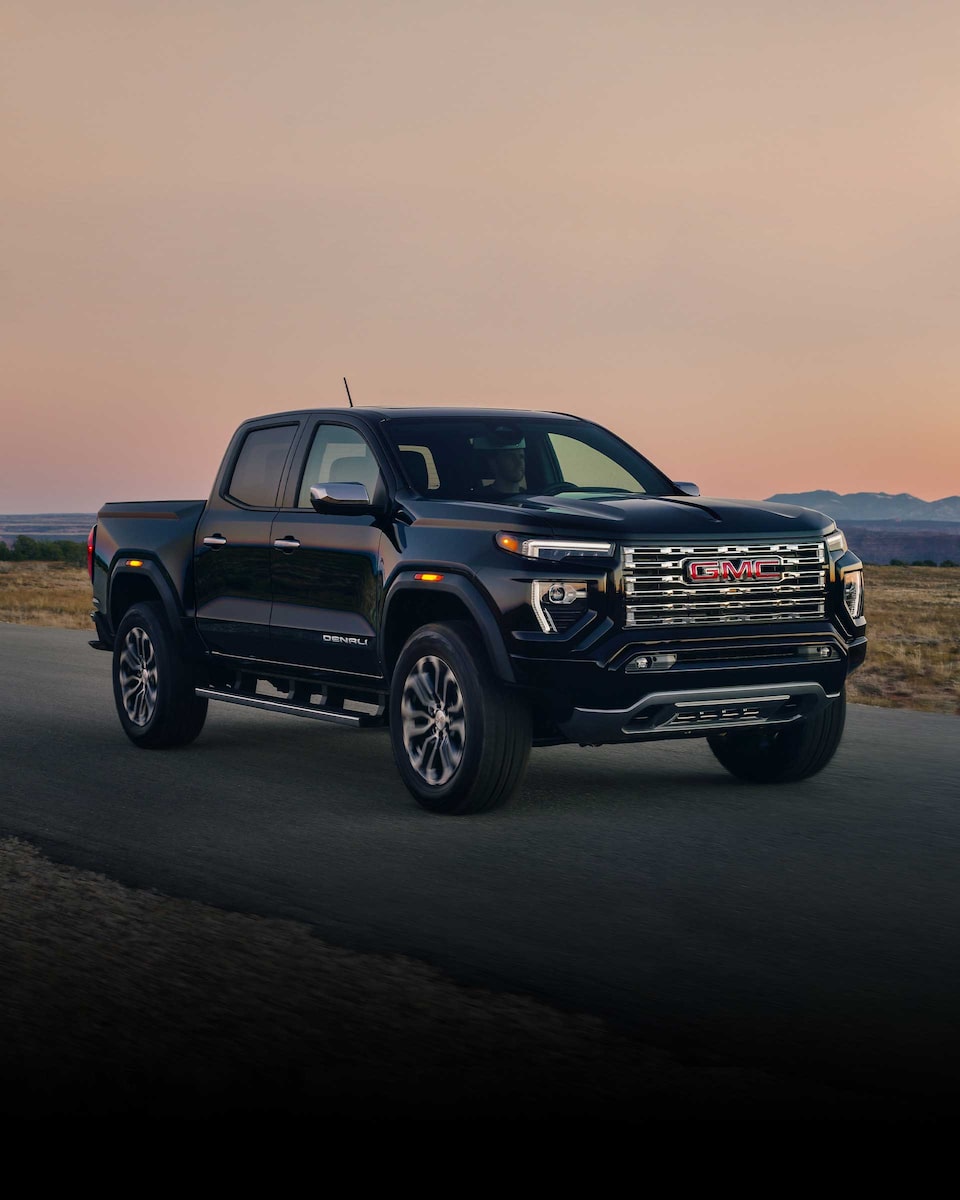 Front Three-Quarters View of a 2024 GMC Canyon Mid-Size Truck Driving on an Open Road at Dusk