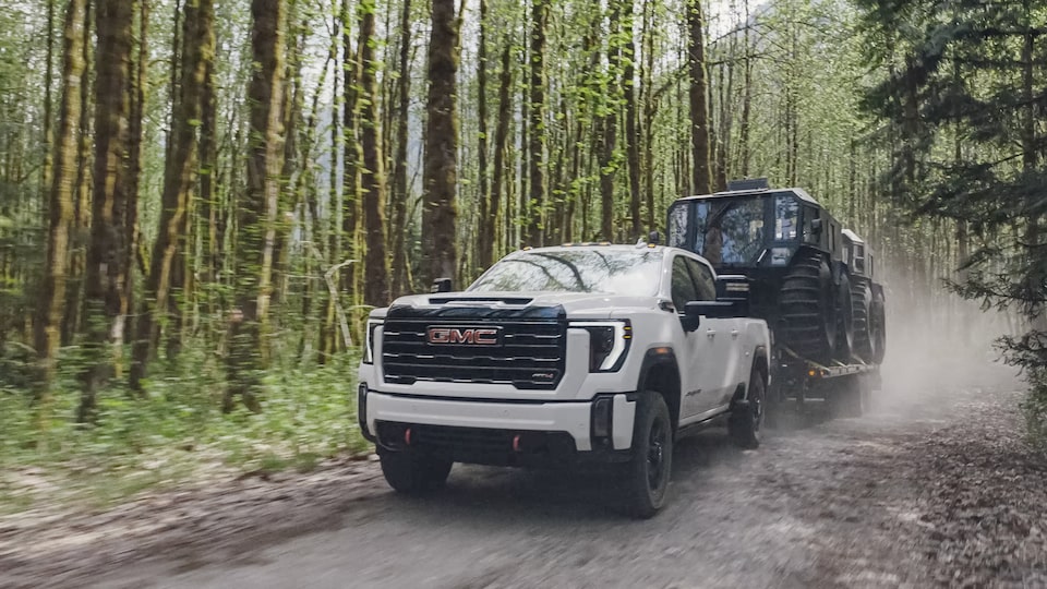 Front Three-Quarters View of a GMC Truck Pulling a Trailer Loaded with Two Big Centaur UTVs on a Dirt Road Surrounded by Forest